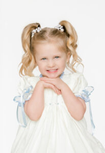 Beautiful toddler blonde girl in an heirloom dress rests her chin on her hands and smiles.