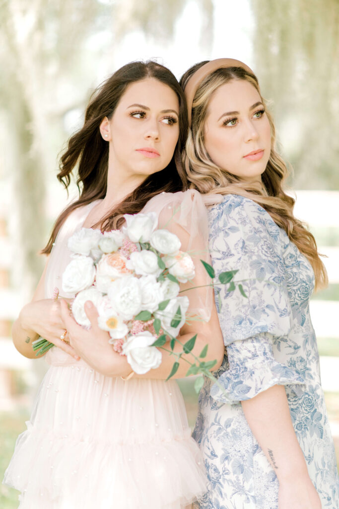 Gorgeous sisters show off their professional hair and makeup standing in front of a white fence holding a bouquet of flowers.
