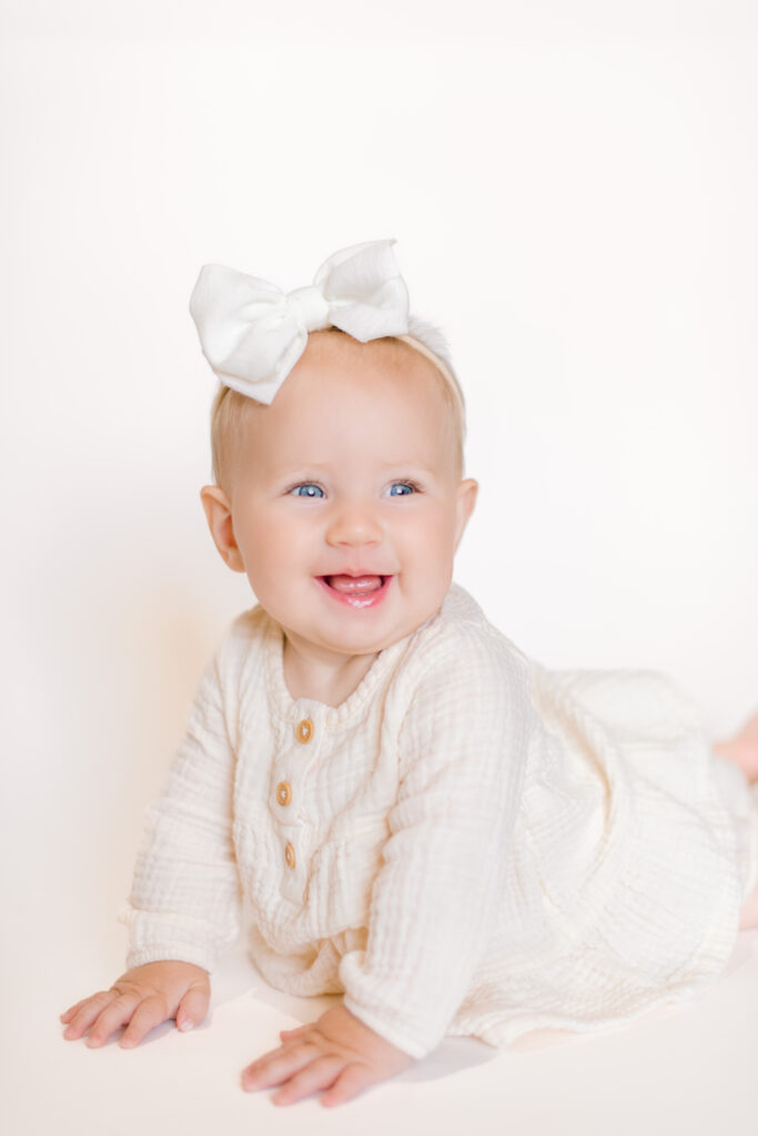 Beautiful baby girl dressed in cream lays on belly holding herself up and smiling.