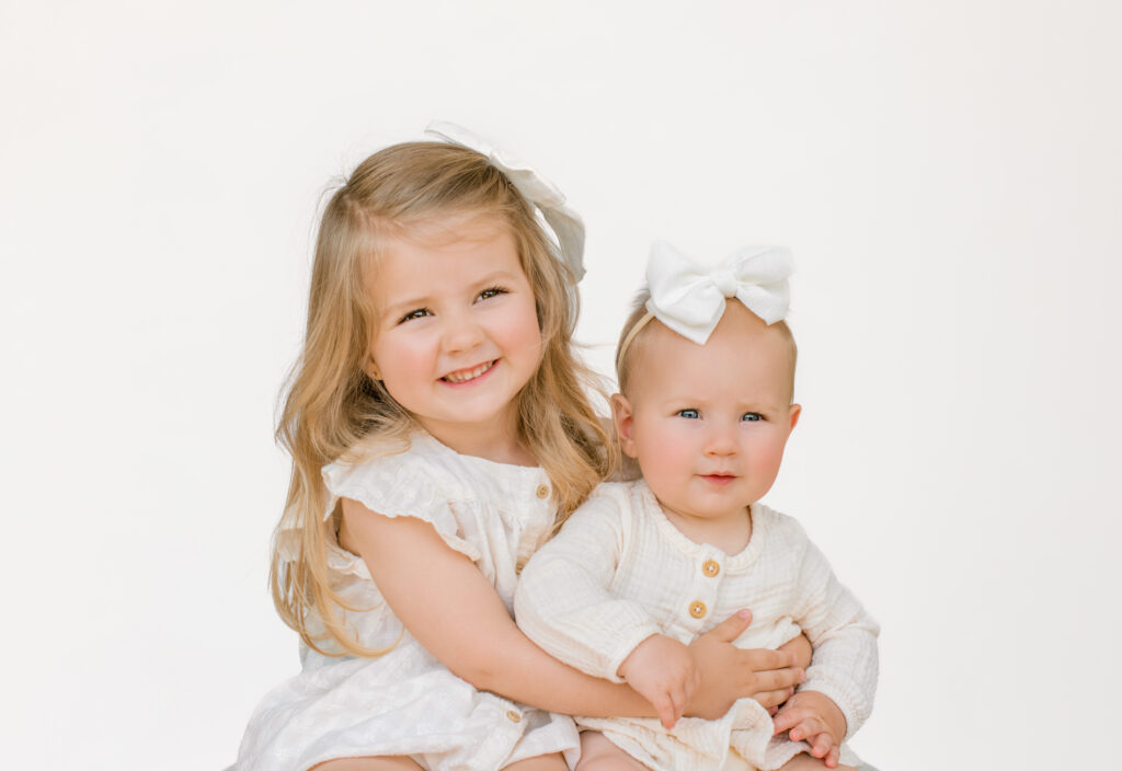 Perfectly posed young sisters sit gracefully dressed in white with a white backdrop.