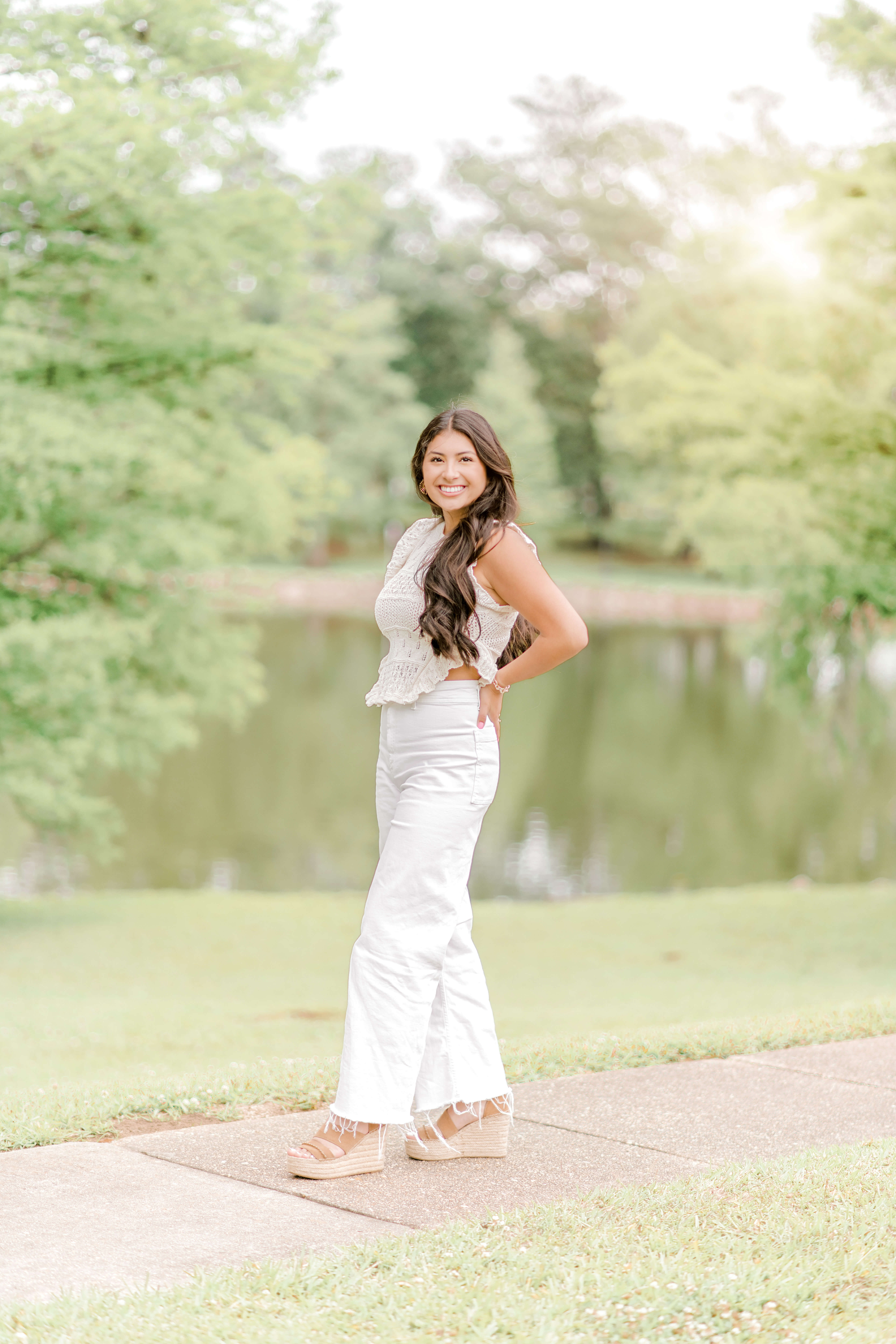 A brunette senior stands in front of a lake with hands on back hips after visiting a coffee shop in Baton Rouge.