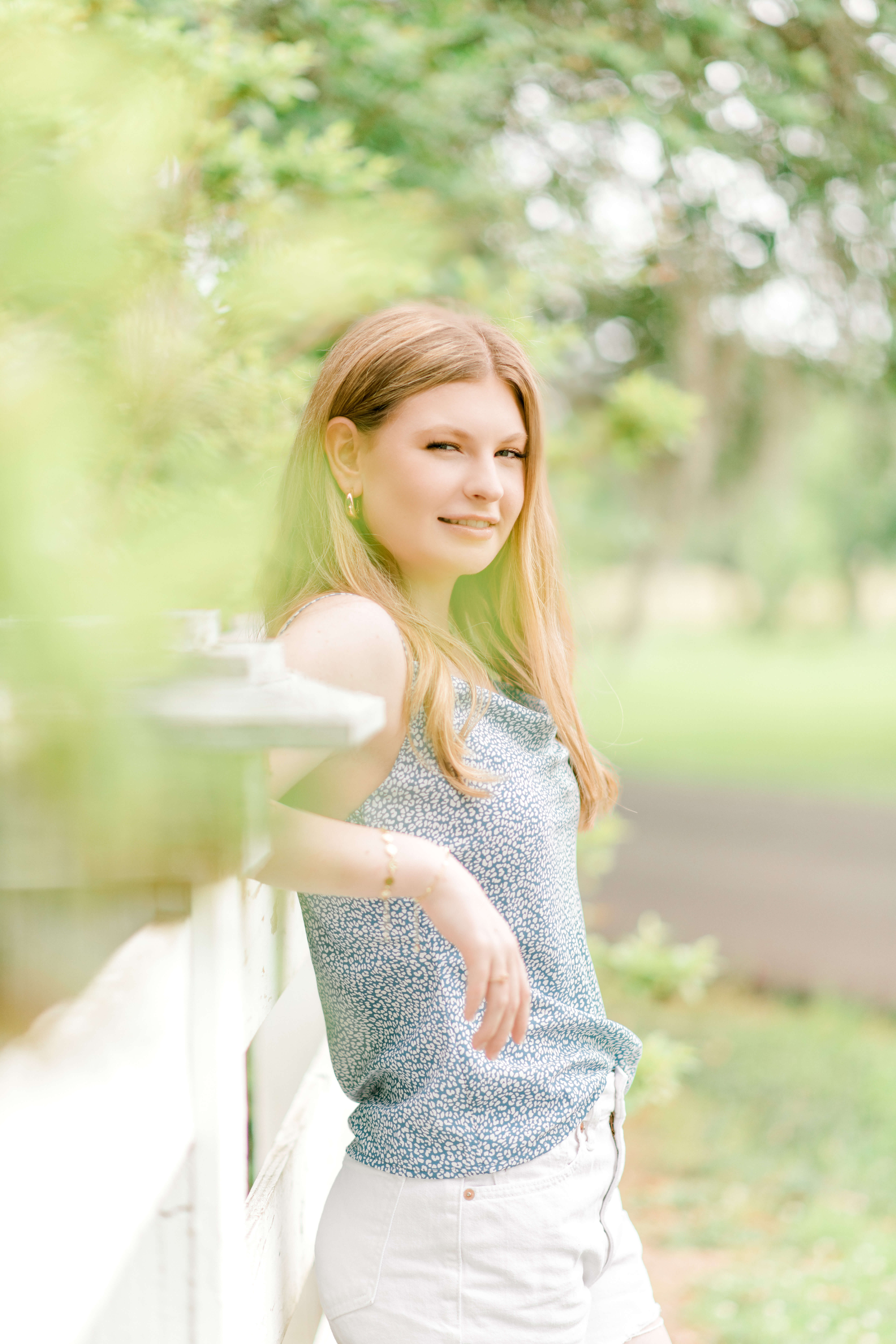 Standing along a white picket fence, a strawberry blonde senior in a silky blue top looks towards her right shoulder and smiles.
