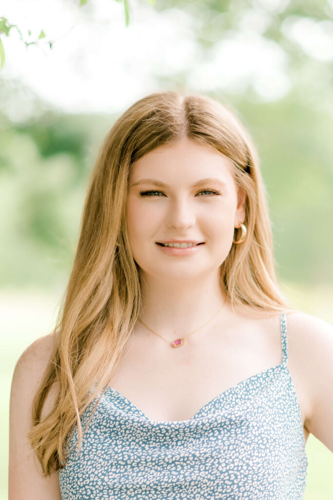 Beautiful senior girl wearing a blue tank top with hair tucked behind left ear stands for a close-up portrait during a Baton Rouge senior session.
