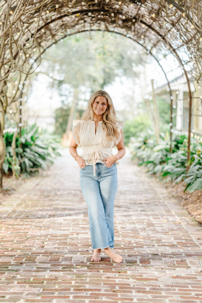 Senior in jeans and ivory top stands in vine tunnel at Houma's House in Darrow, LA.