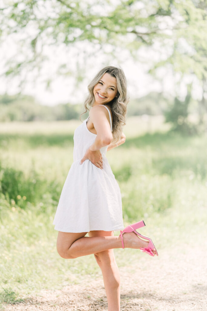 Senior girl in a white dress kicks back her left foot to show off her pink heels in a field in Baton Rouge, LA.