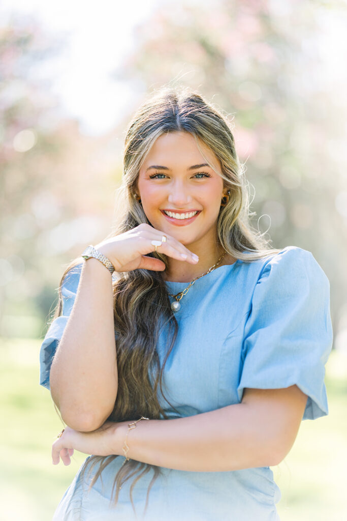 Gorgeous girl in blue dress lightly rests her chin on her relaxed hand with a big smile for her senior portraits.