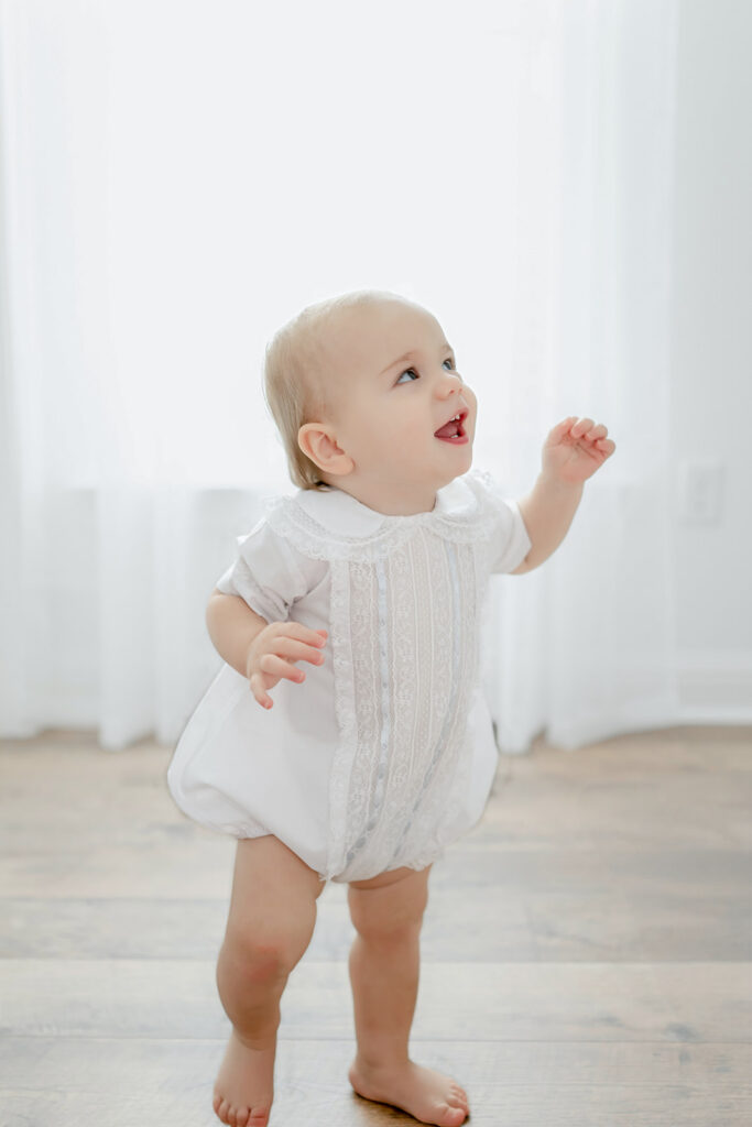 In an outfit from Olly-Olly Children's Store Baton Rouge, a young child walks in an all white room in front of a window for a first birthday session in Baton Rouge, LA.