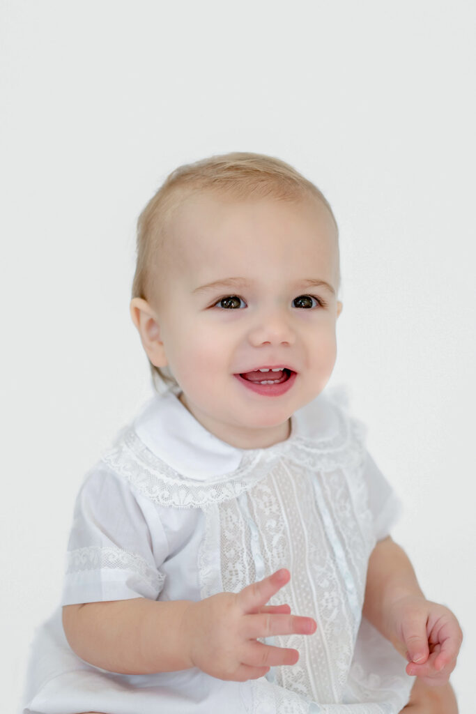 Toddler sits in front a white backdrop and smiles for heirloom photos with Morgan Leigh Photography wearing clothing from Olly-Olly Children's Store Baton Rouge.