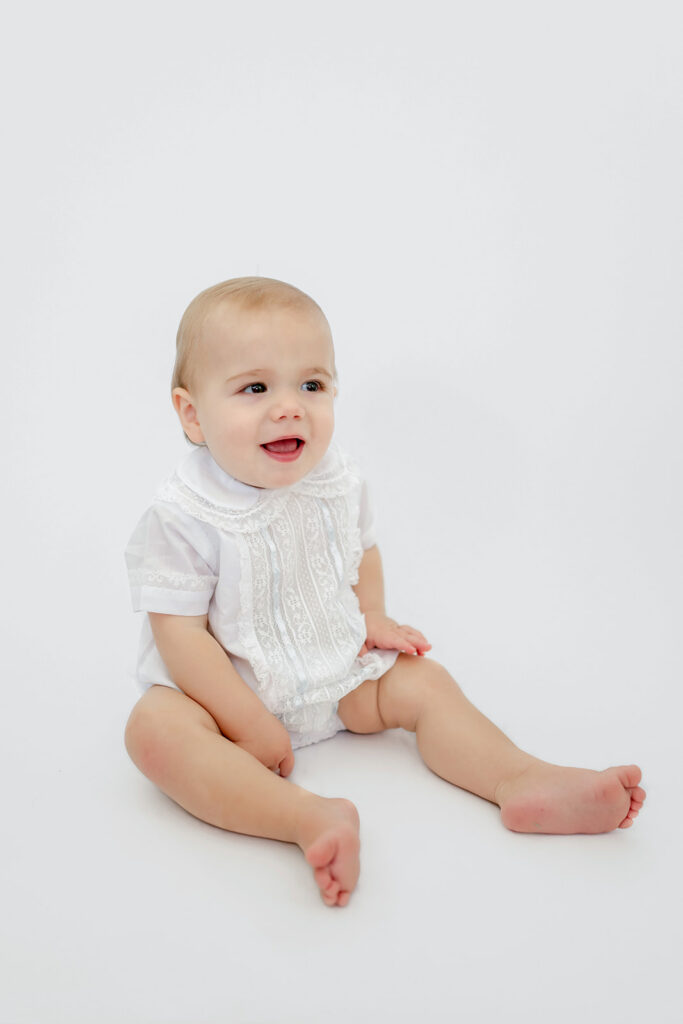 Toddler in heirloom outfit sits on white backdrop for a session in Denham Springs, LA.