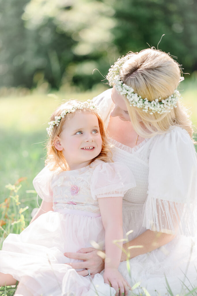 Young girl sits in mom's lap and looks back at her wearing flower crowns during a session with Morgan Leigh Photography.