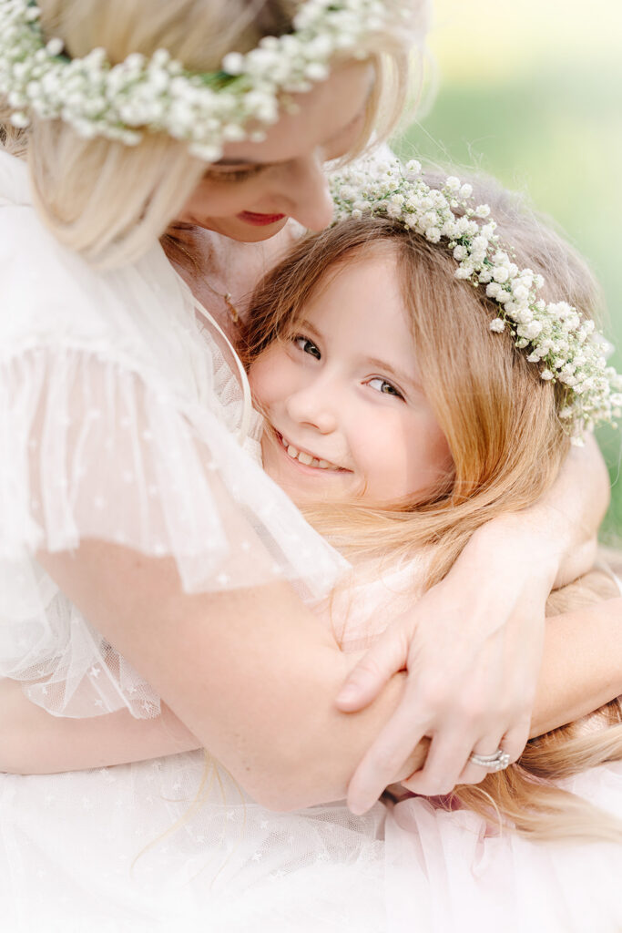 Young daughter embraces mom wearing light dresses and flower crowns in a Baton Rouge field.