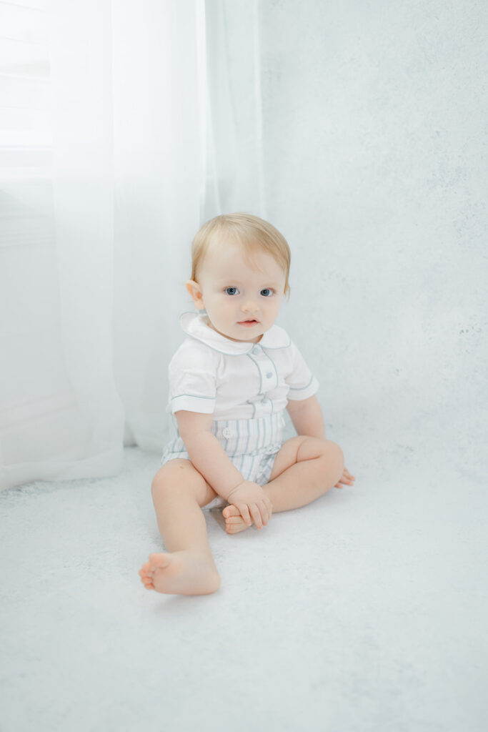 Baby in blue and white outfit sits in front of a window.