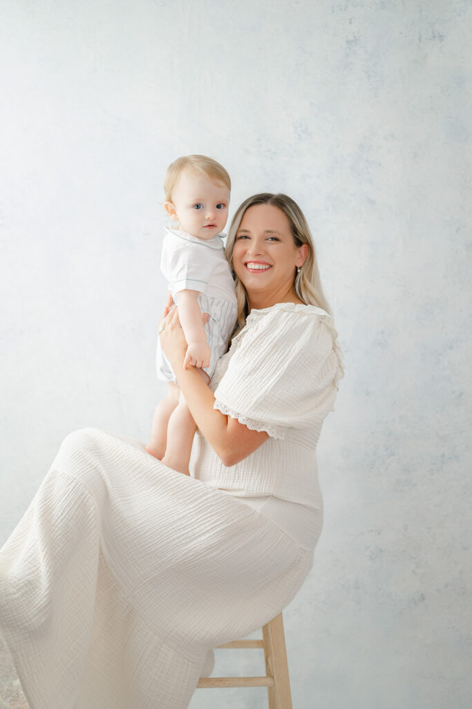 Mother sits on stool in front of a light blue hand painted backdrop holding her toddler son.