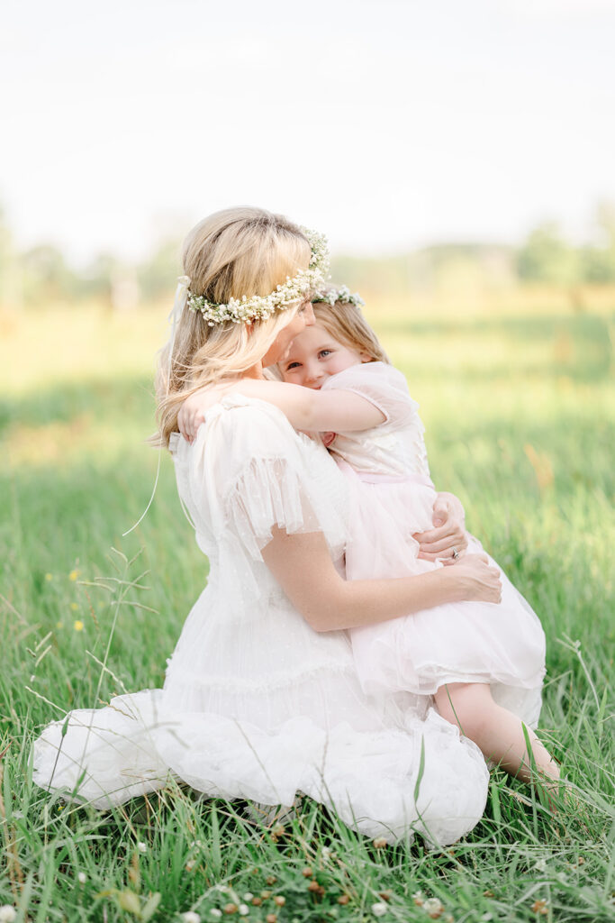 Mother and daughter embrace whilst sitting in a field for photos.